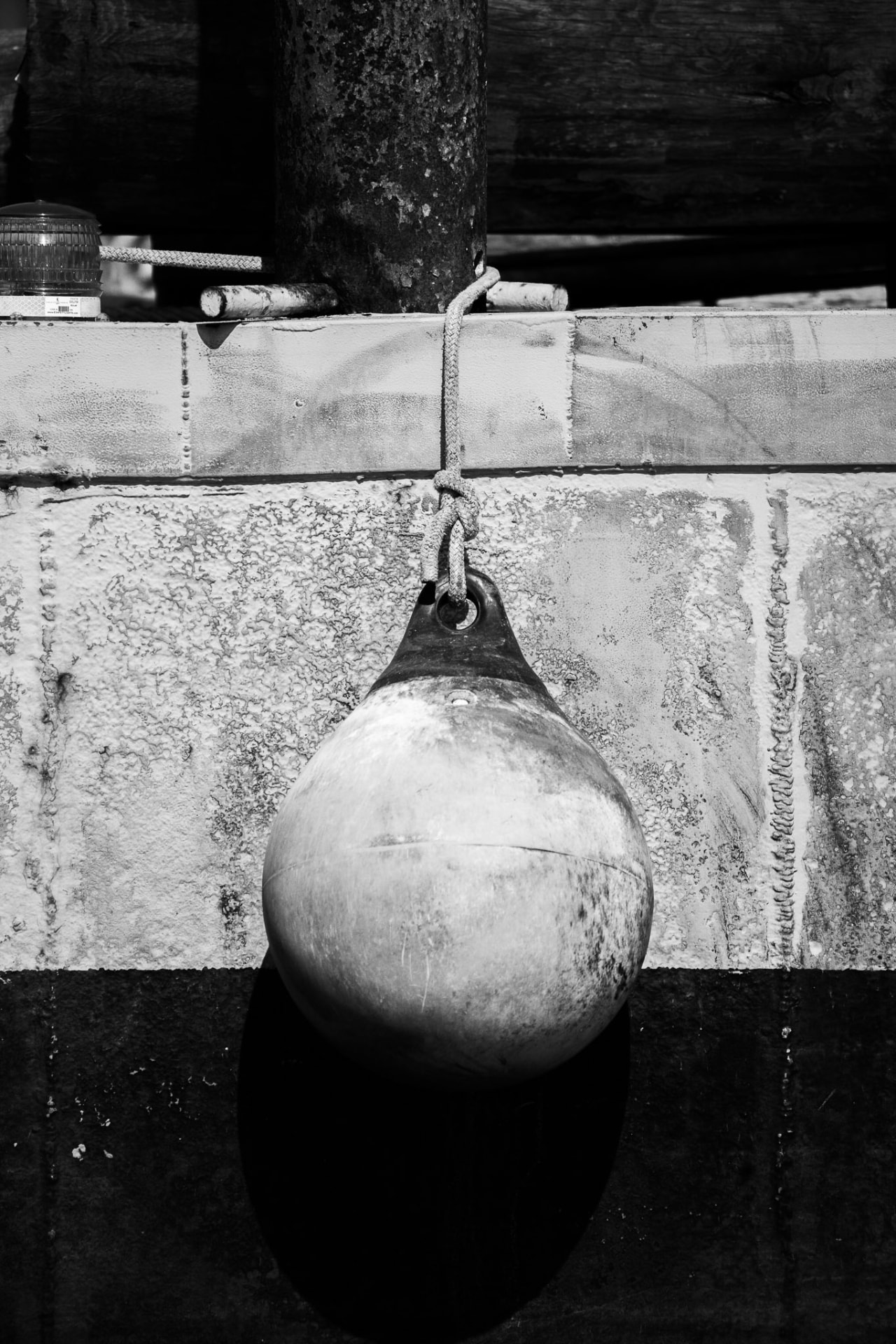 A weathered mooring buoy hanging against a concrete seawall, textured rope and maritime hardware in black and white