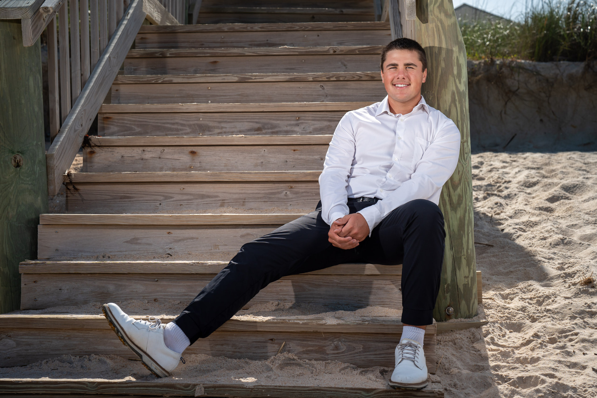 Senior portrait of a young man in a white dress shirt sitting on weathered wooden beach stairs in the sand