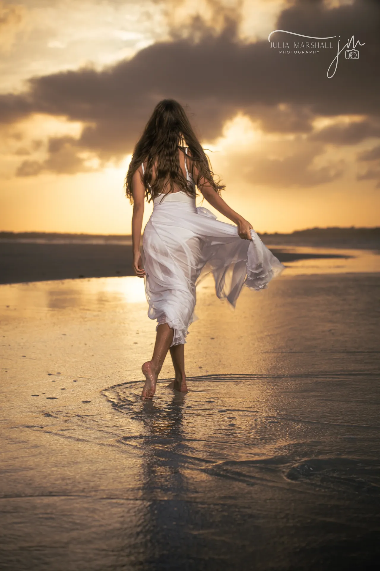 Woman in a flowing white dress walking through the wet sand at sunrise, gold light reflecting around her