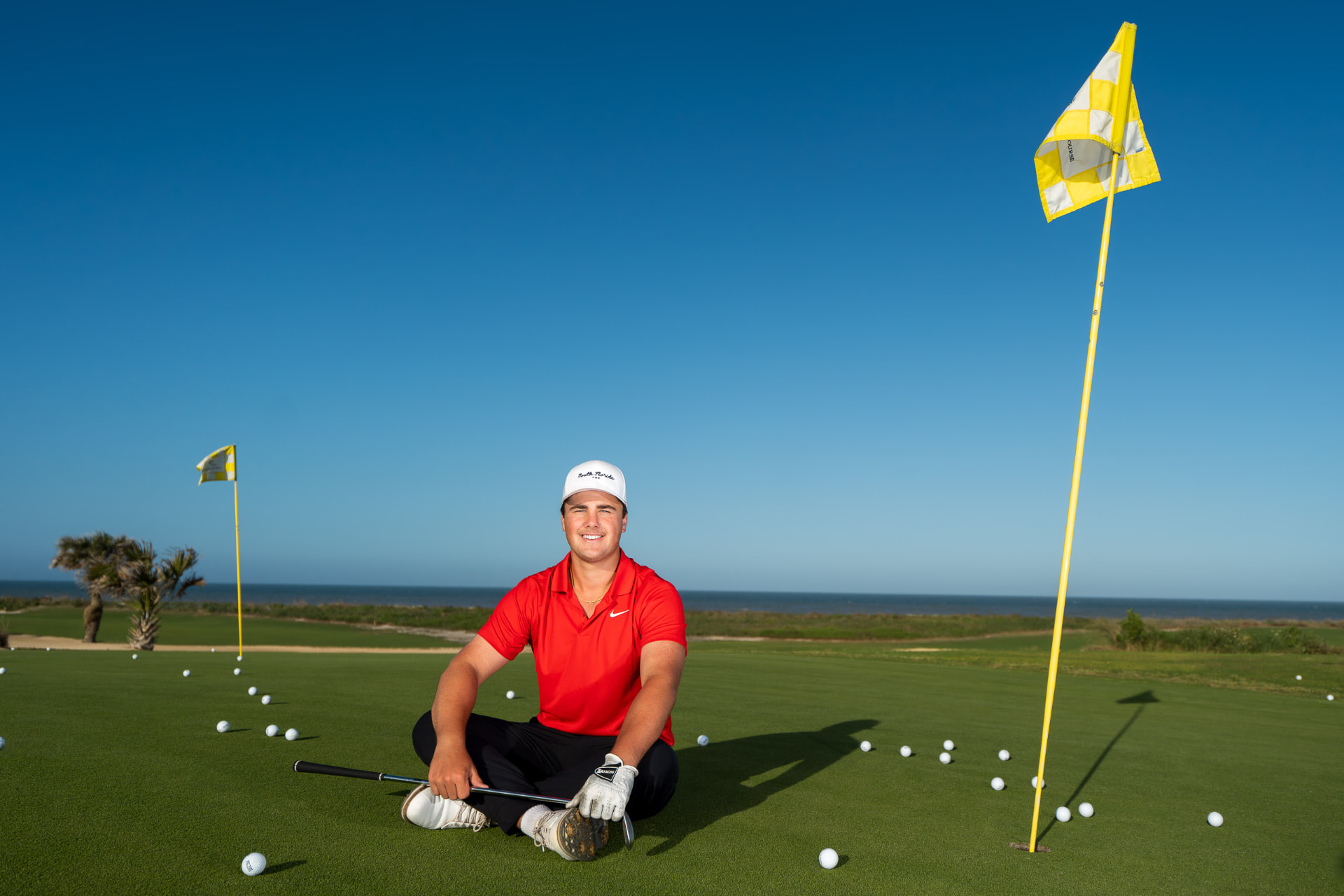Senior portrait of a young man in a red golf polo sitting cross-legged on a putting green surrounded by golf balls beneath a deep blue sky