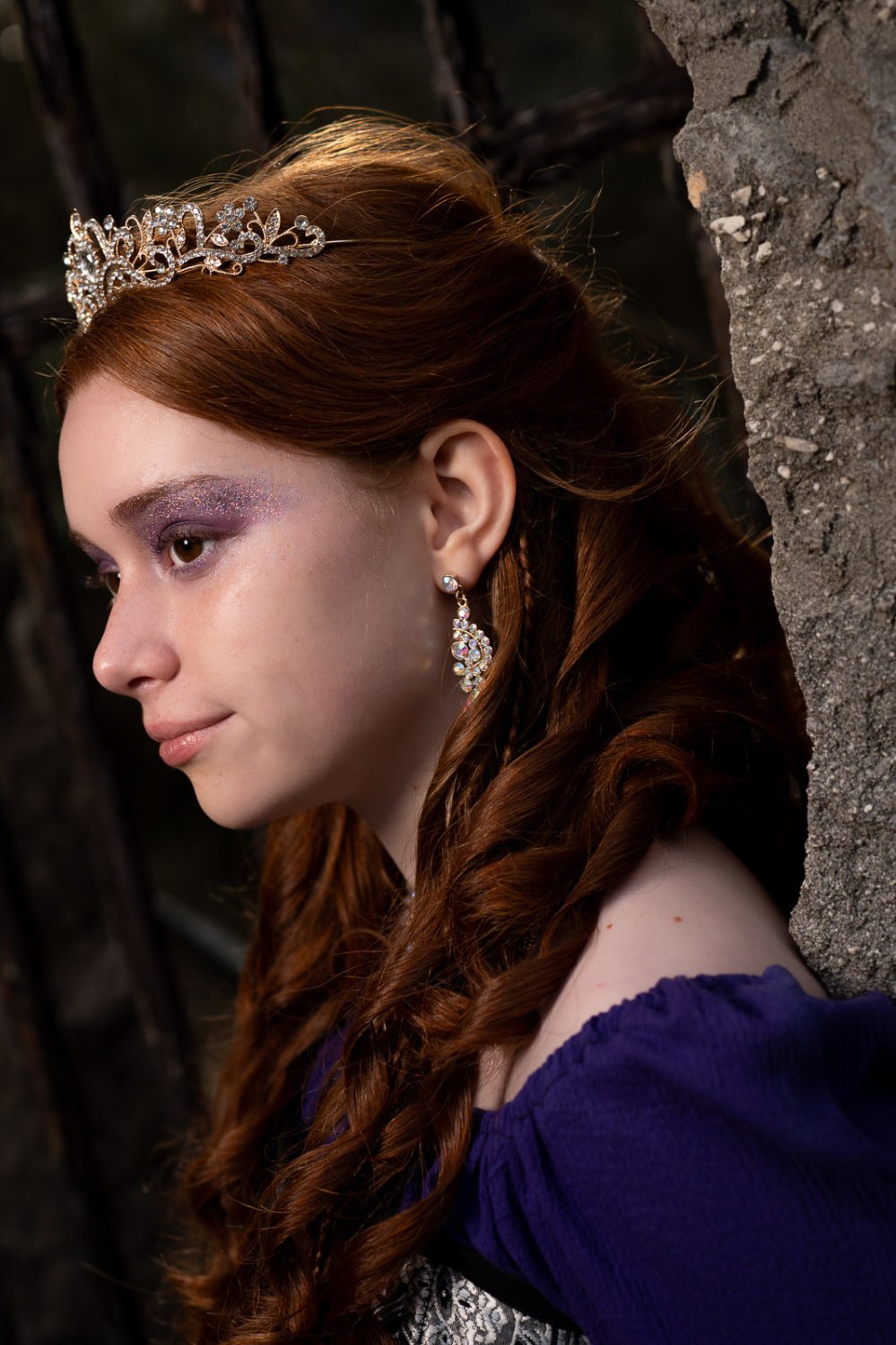Profile portrait of a young woman with auburn braids, a jeweled tiara and shimmering violet makeup resting against a stone wall