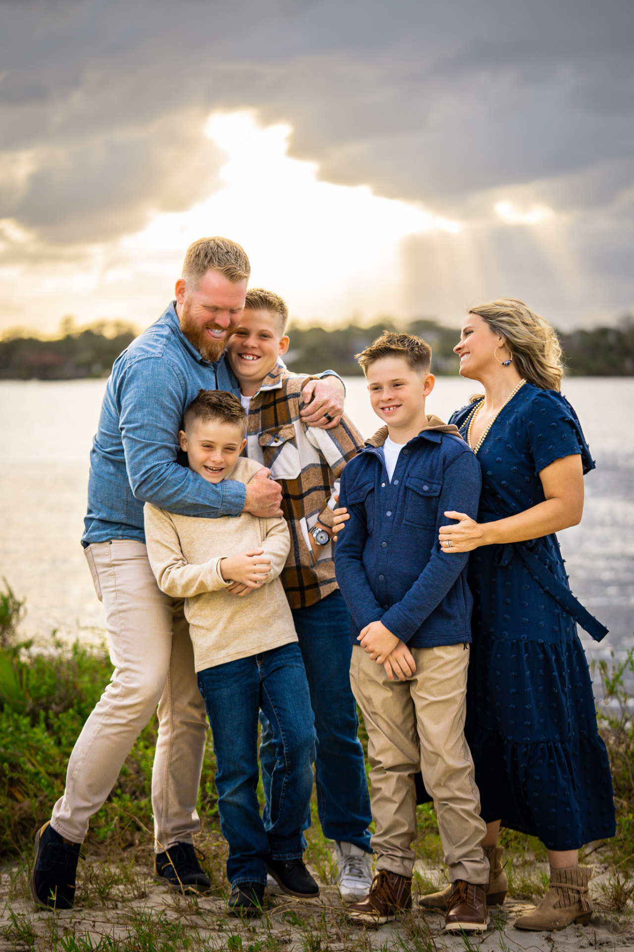 Family of five laughing together at the edge of a lake as golden sunset light breaks through dramatic clouds