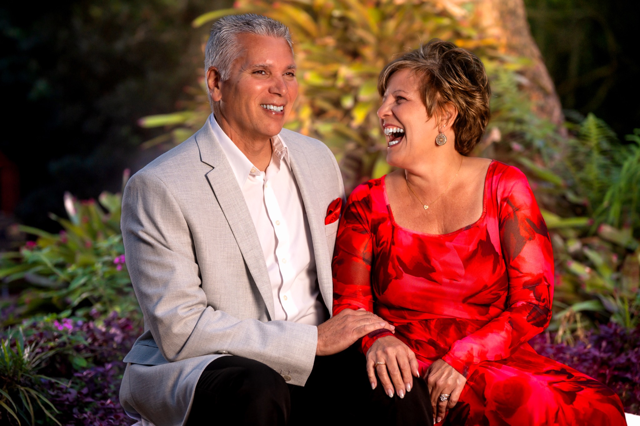 Couple laughing together while seated in a sunlit tropical garden, the woman in a red floral dress