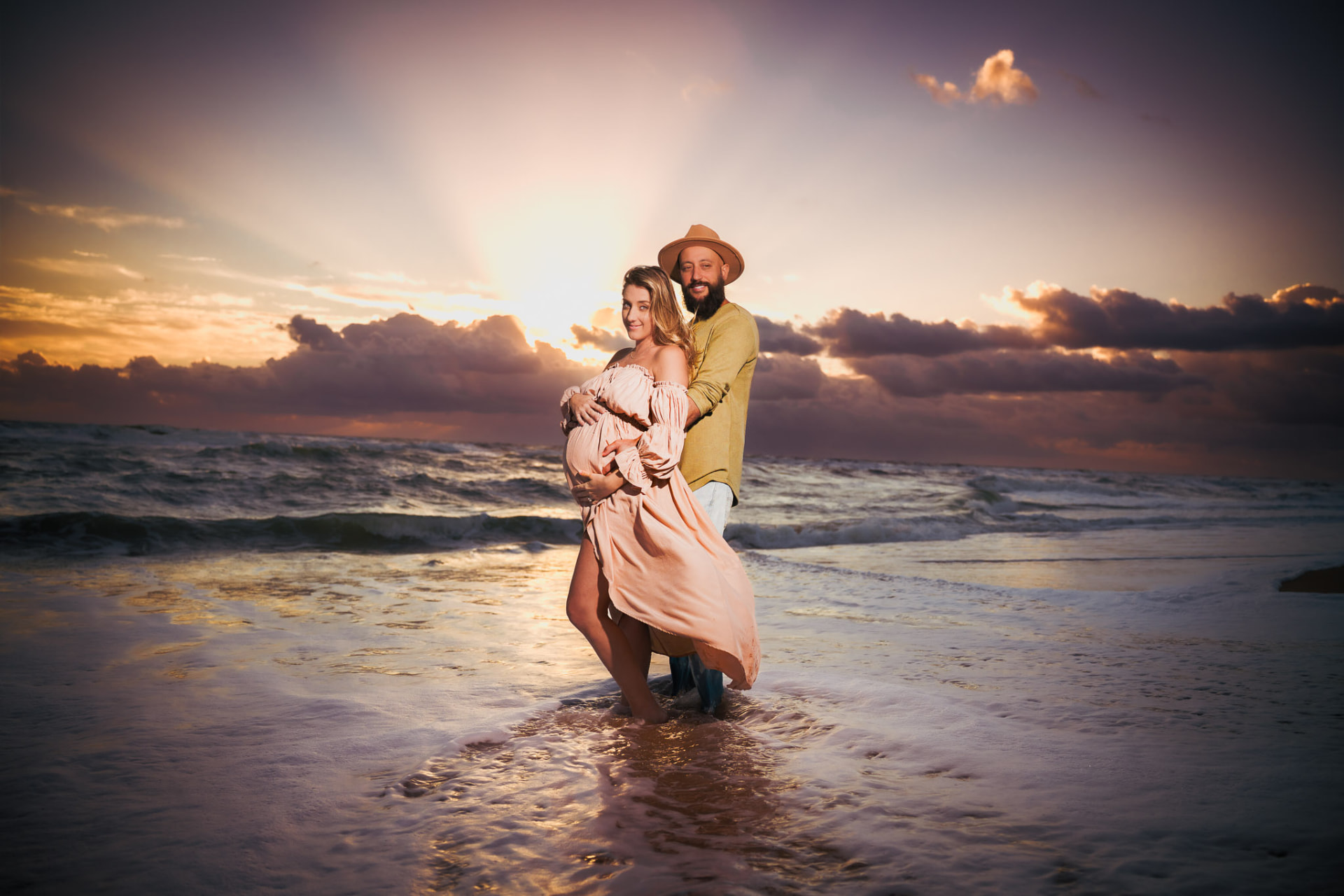 Expectant couple embracing at sunset on the beach, the woman in a flowing peach off-shoulder dress and the man in a mustard shirt and hat