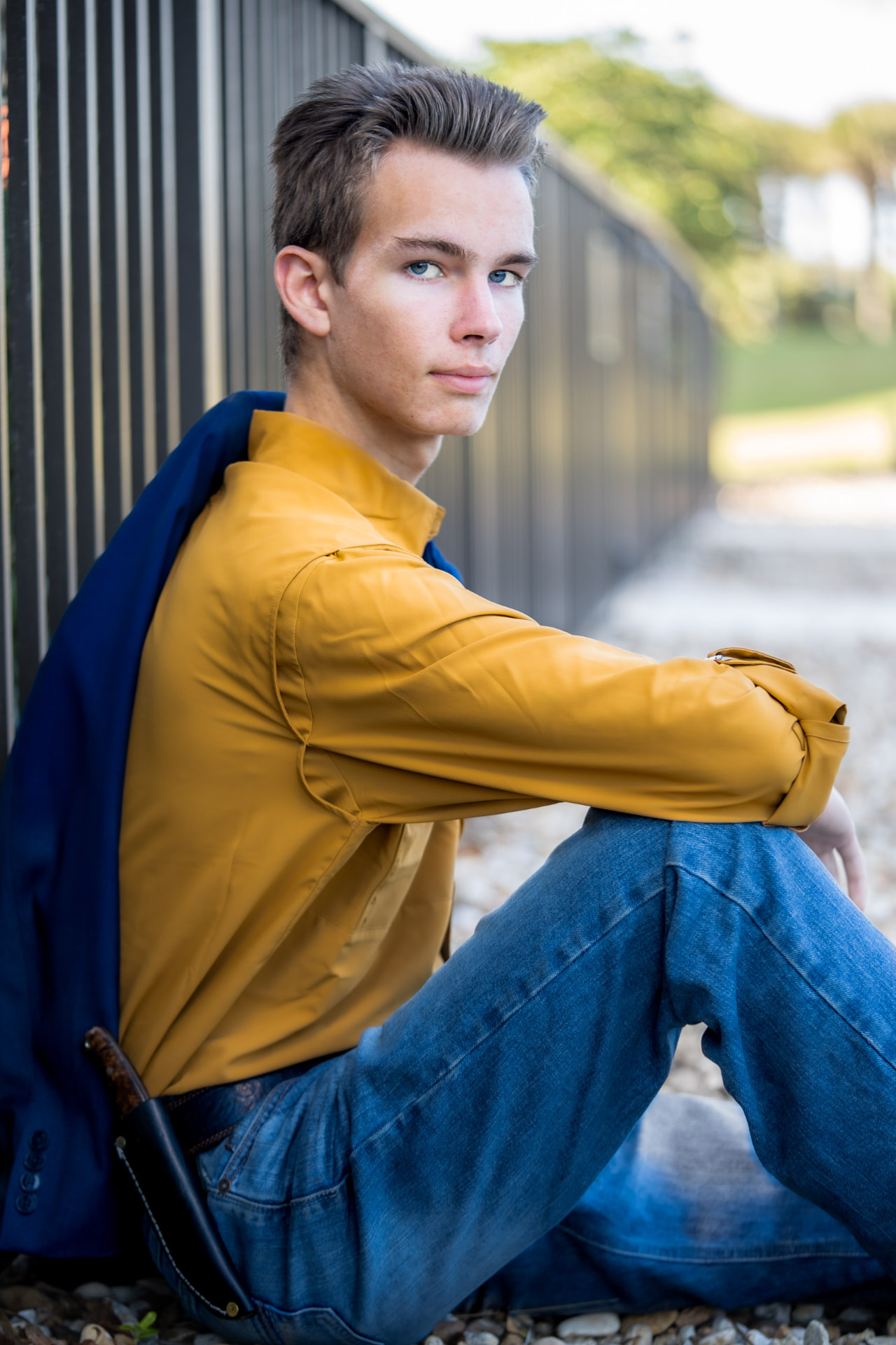 Senior portrait of a young man in a mustard shirt and jeans seated against a dark metal fence with a navy jacket over his shoulders