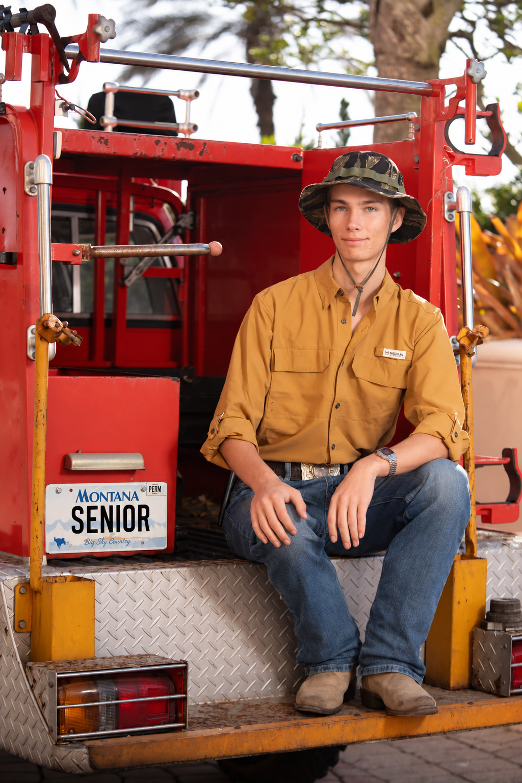Senior portrait of a young man in a camo bucket hat and work shirt seated on a vintage red fire truck beside a Montana senior license plate