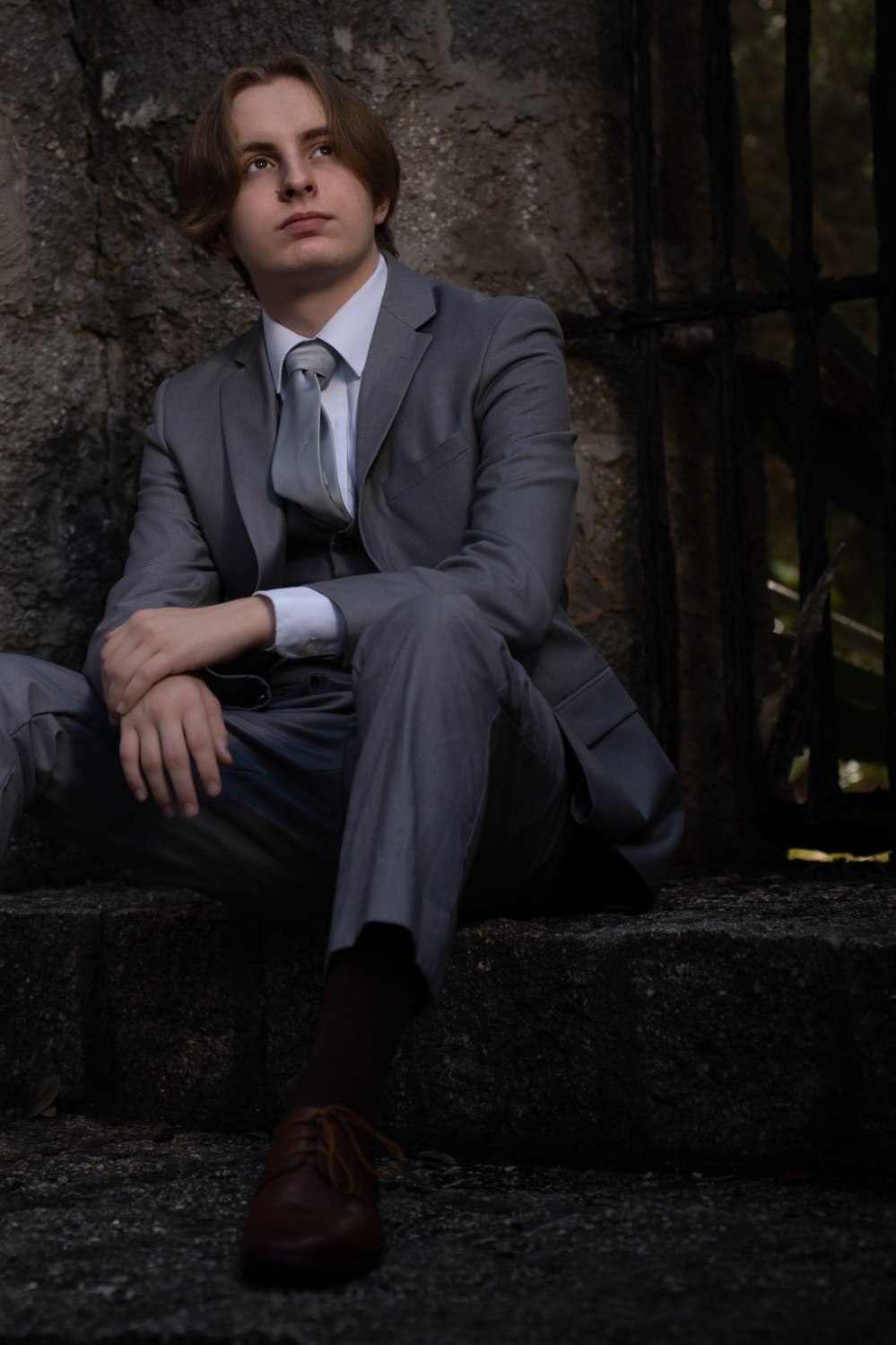 Senior portrait of a young man in a grey three-piece suit seated on stone steps against a coquina wall
