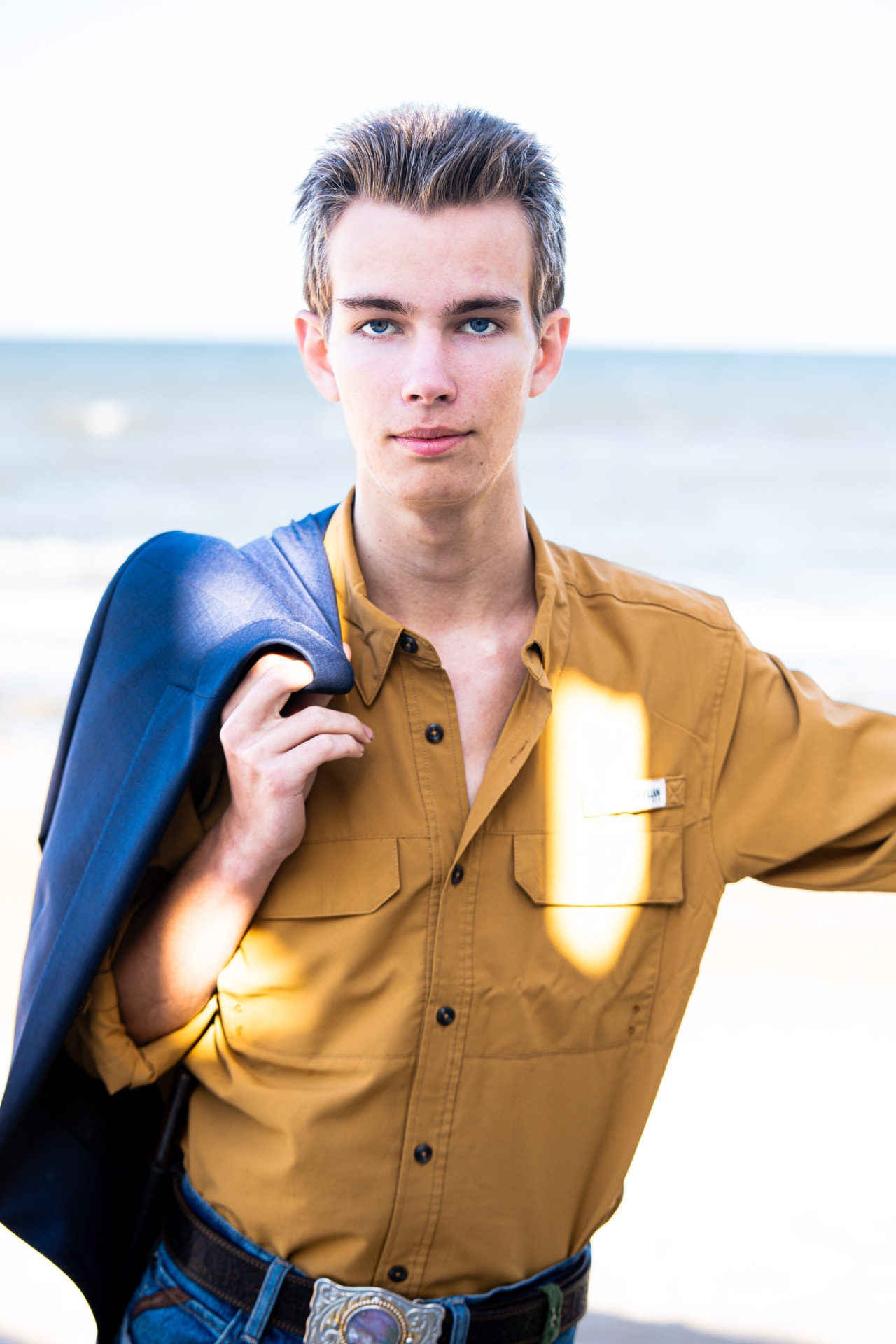 Senior portrait of a young man at the beach with a navy jacket slung over his shoulder in bright backlight