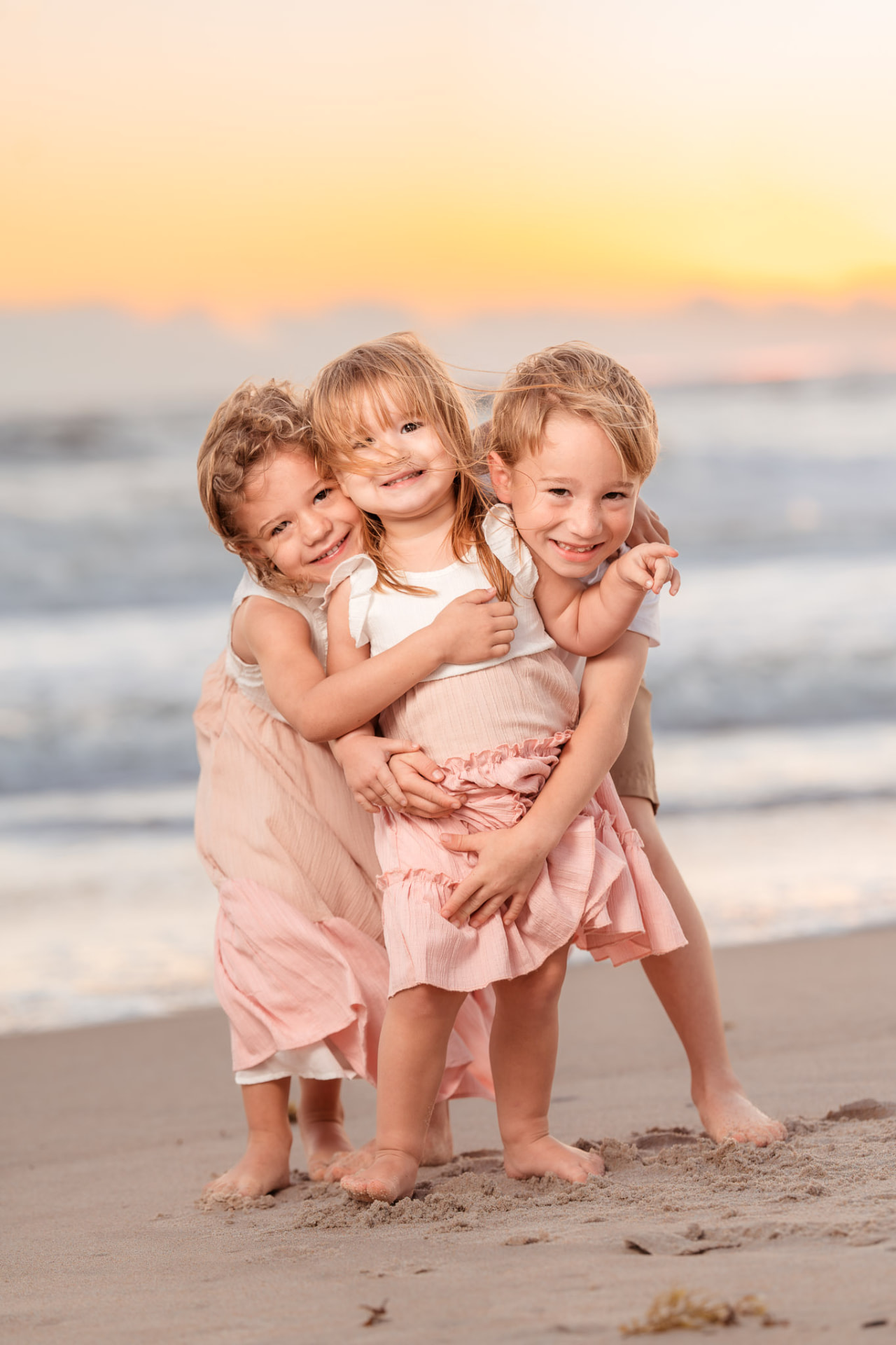 Three young siblings in soft pink outfits hugging and laughing on the beach at sunset