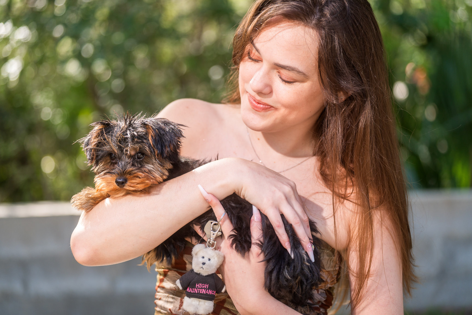 Young woman holding a small Yorkie puppy with a tiny teddy bear charm in sunny greenery