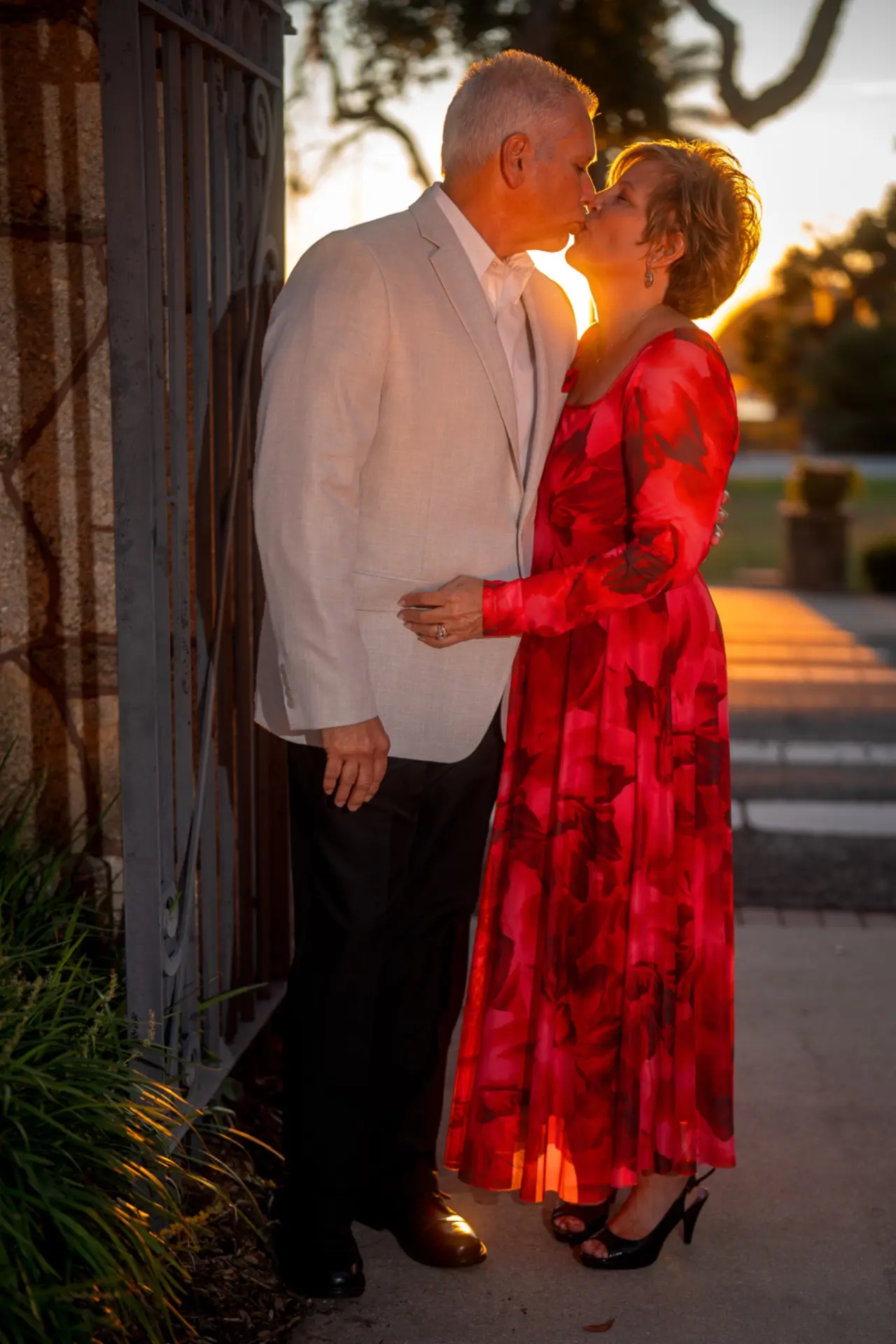 Couple sharing a kiss beside an iron garden gate as golden sunset light glows behind them, the woman in a long red floral gown
