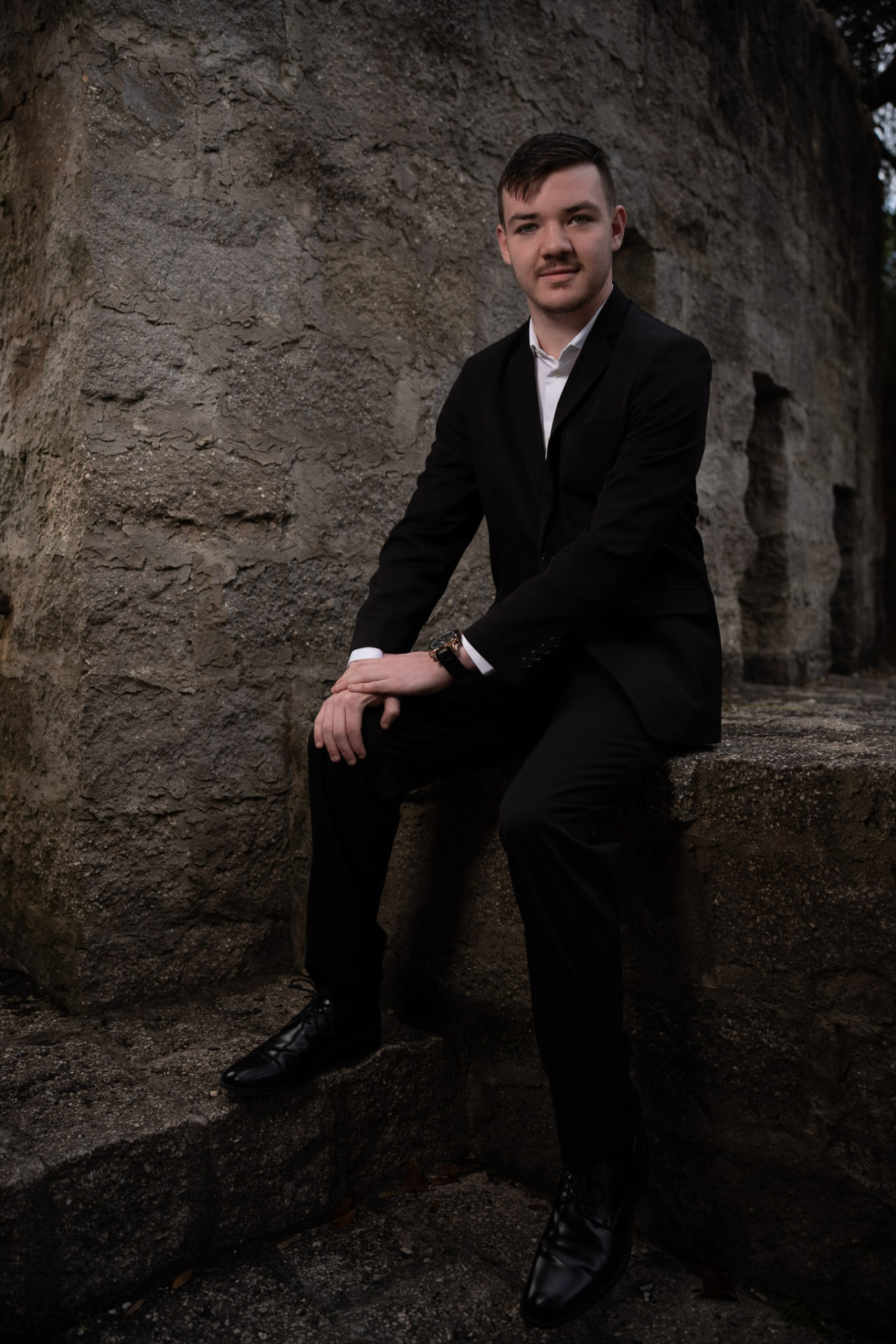 Young man in a black suit seated on weathered stone steps against a textured wall