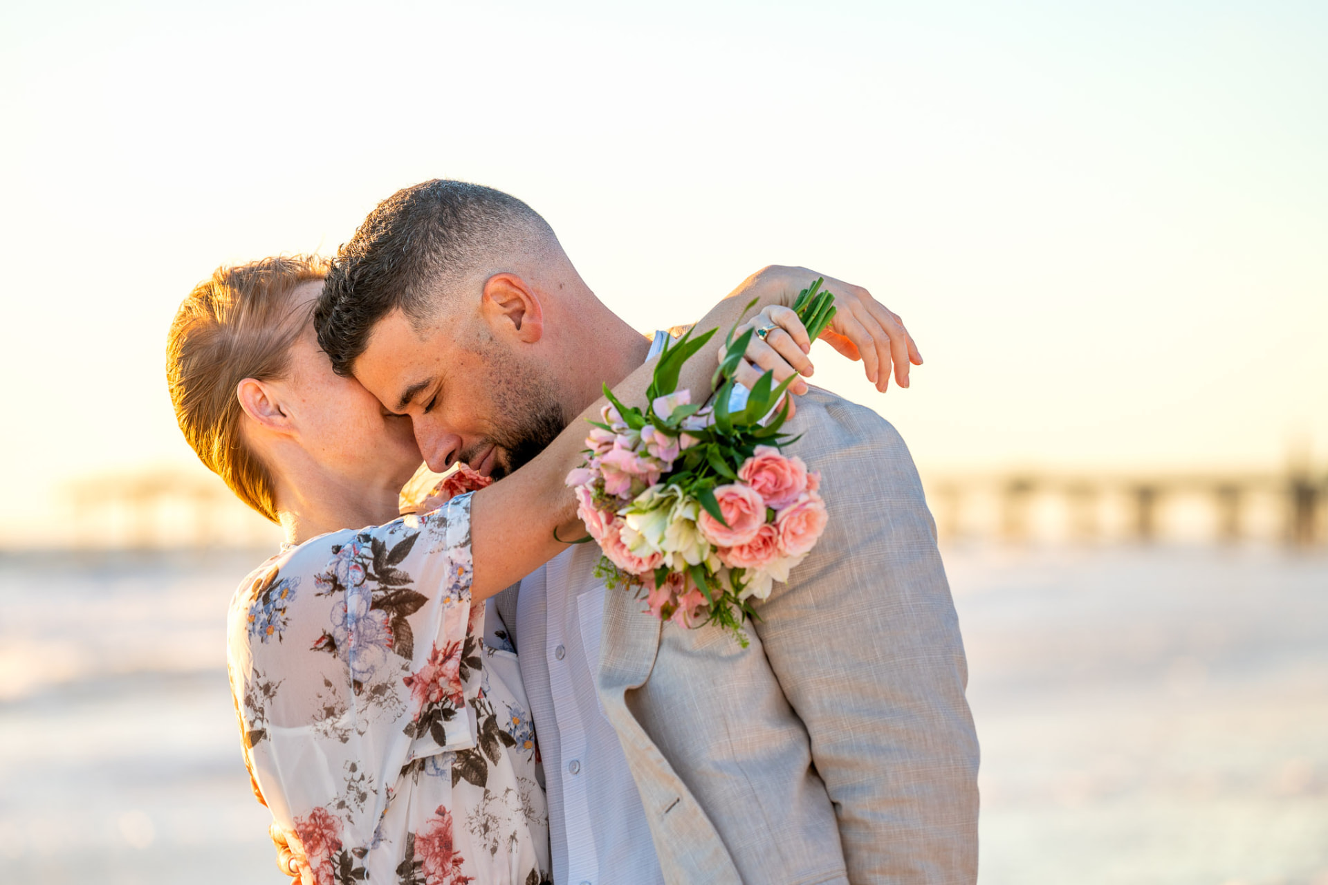 Couple embracing on a sunlit beach, the woman in a flowing floral dress holding a pink bouquet around the man's shoulders