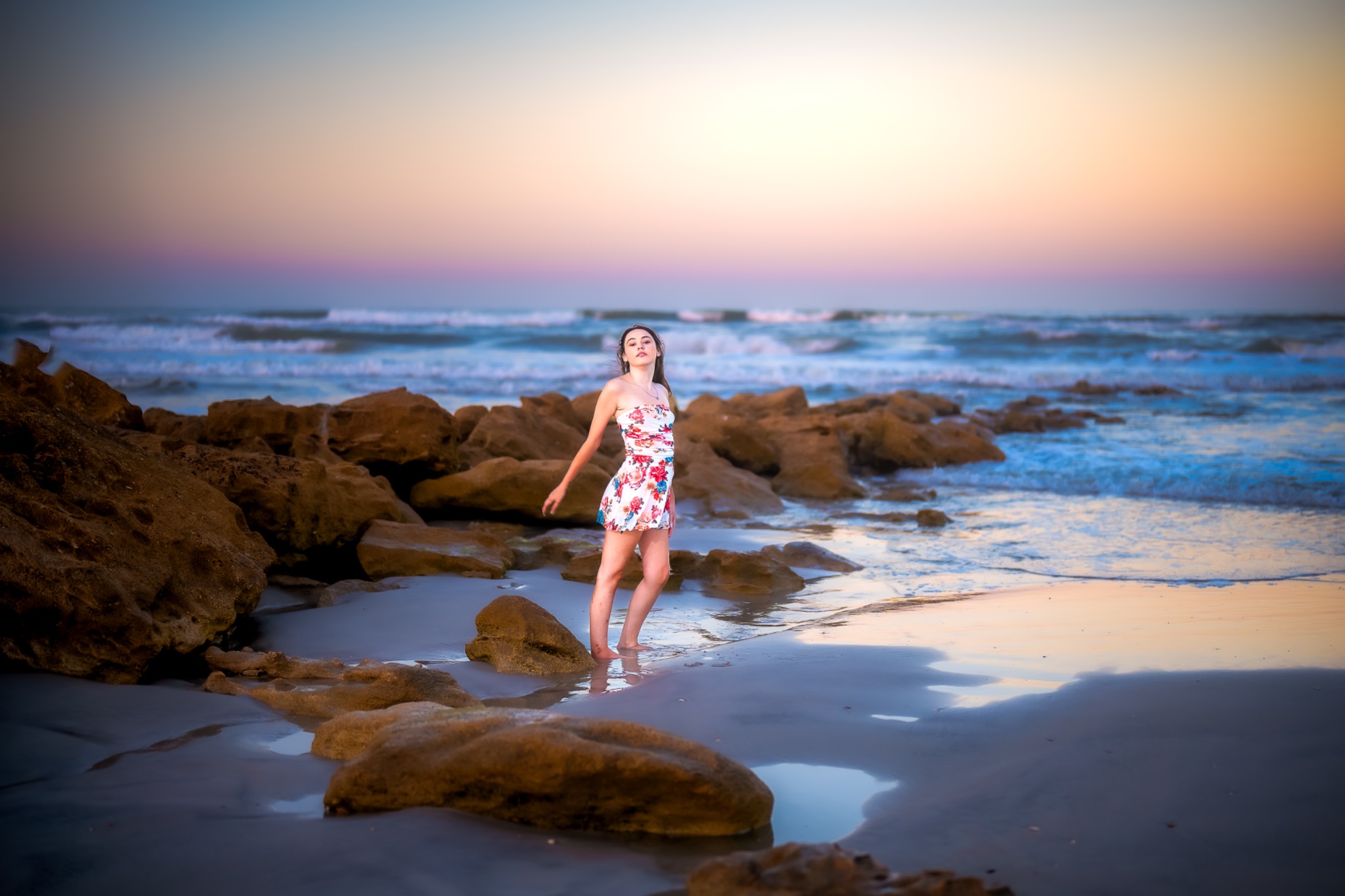 Young woman in a floral dress standing among coquina rocks at the ocean's edge as pastel dusk light spreads across the surf