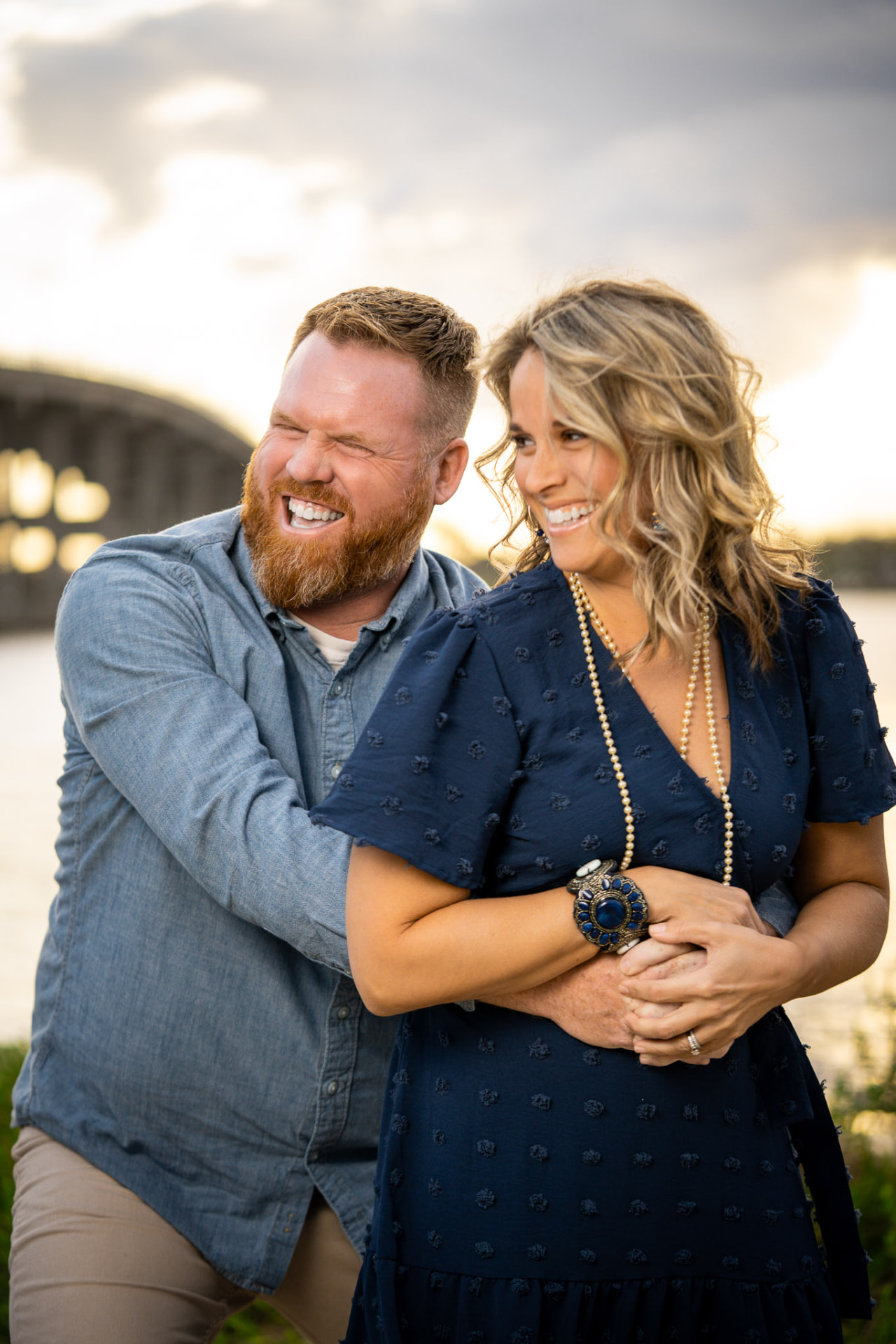 Couple embracing and laughing together near a coastal bridge at sunset