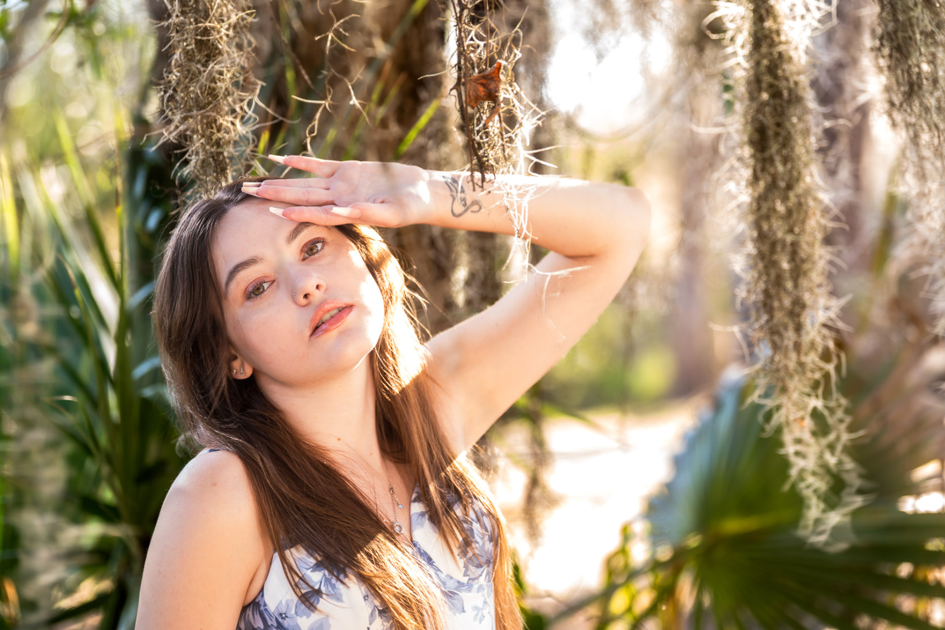 Young woman in a floral dress shielding her eyes from golden sunlight among Spanish moss