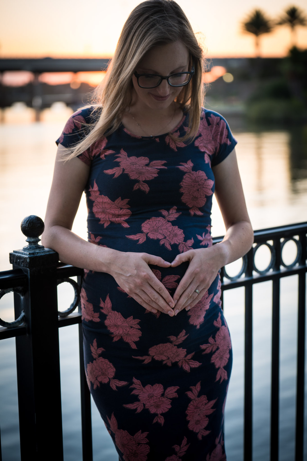 Expectant mother in a navy floral dress forming a heart with her hands over her baby bump by a waterfront railing at sunset