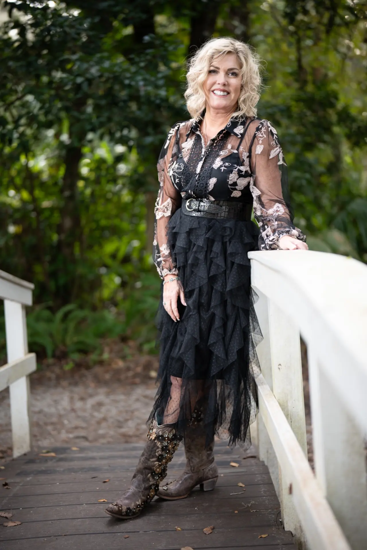 Woman in a tiered black dress and boots smiling on a wooden footbridge surrounded by lush greenery