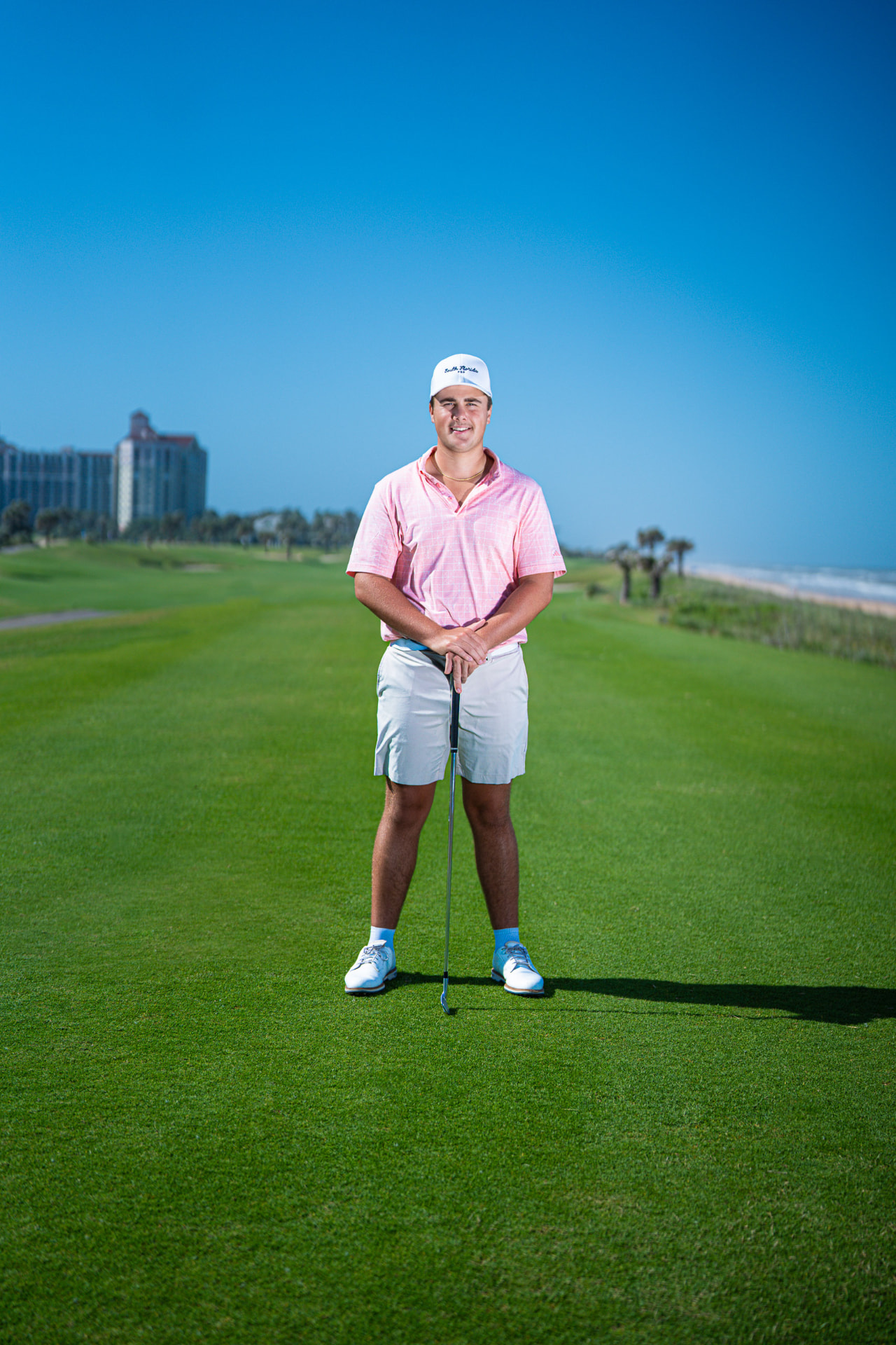 Senior portrait of a young man in a pink golf polo standing on an oceanfront fairway leaning on his club