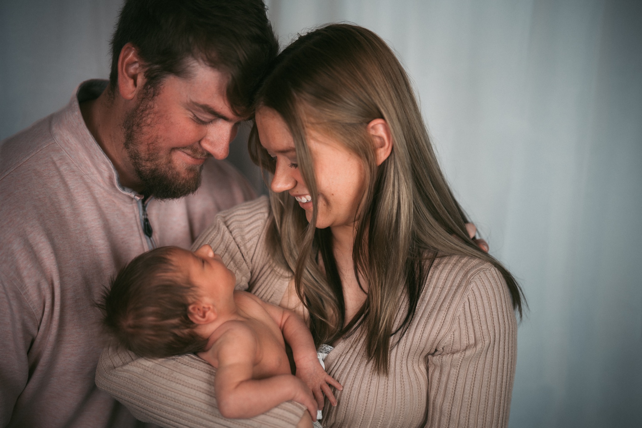 New parents leaning their foreheads together while cradling their sleeping newborn in soft window light