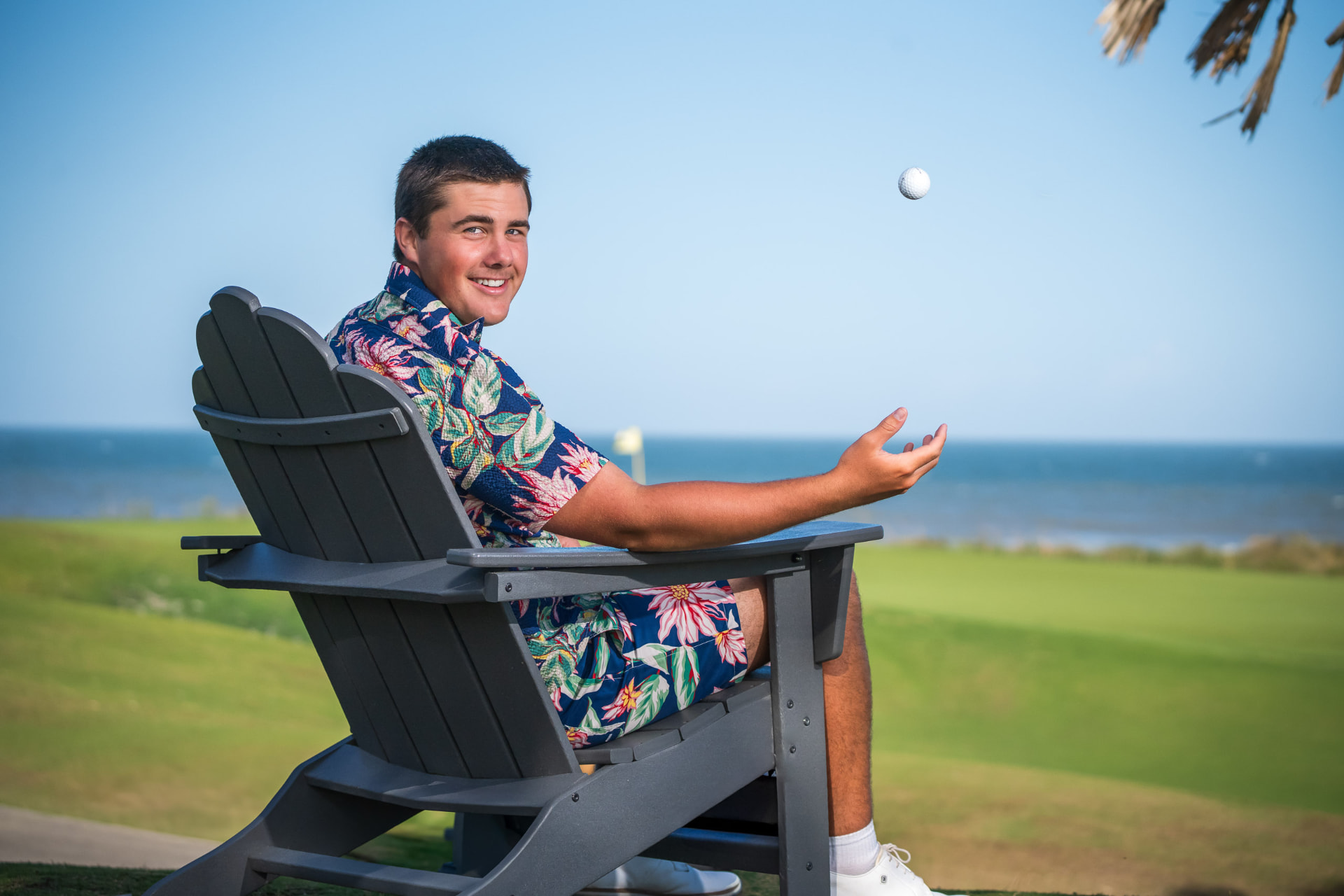 Senior portrait of a young man in a floral shirt relaxing in an Adirondack chair beside the ocean, tossing a golf ball into the air