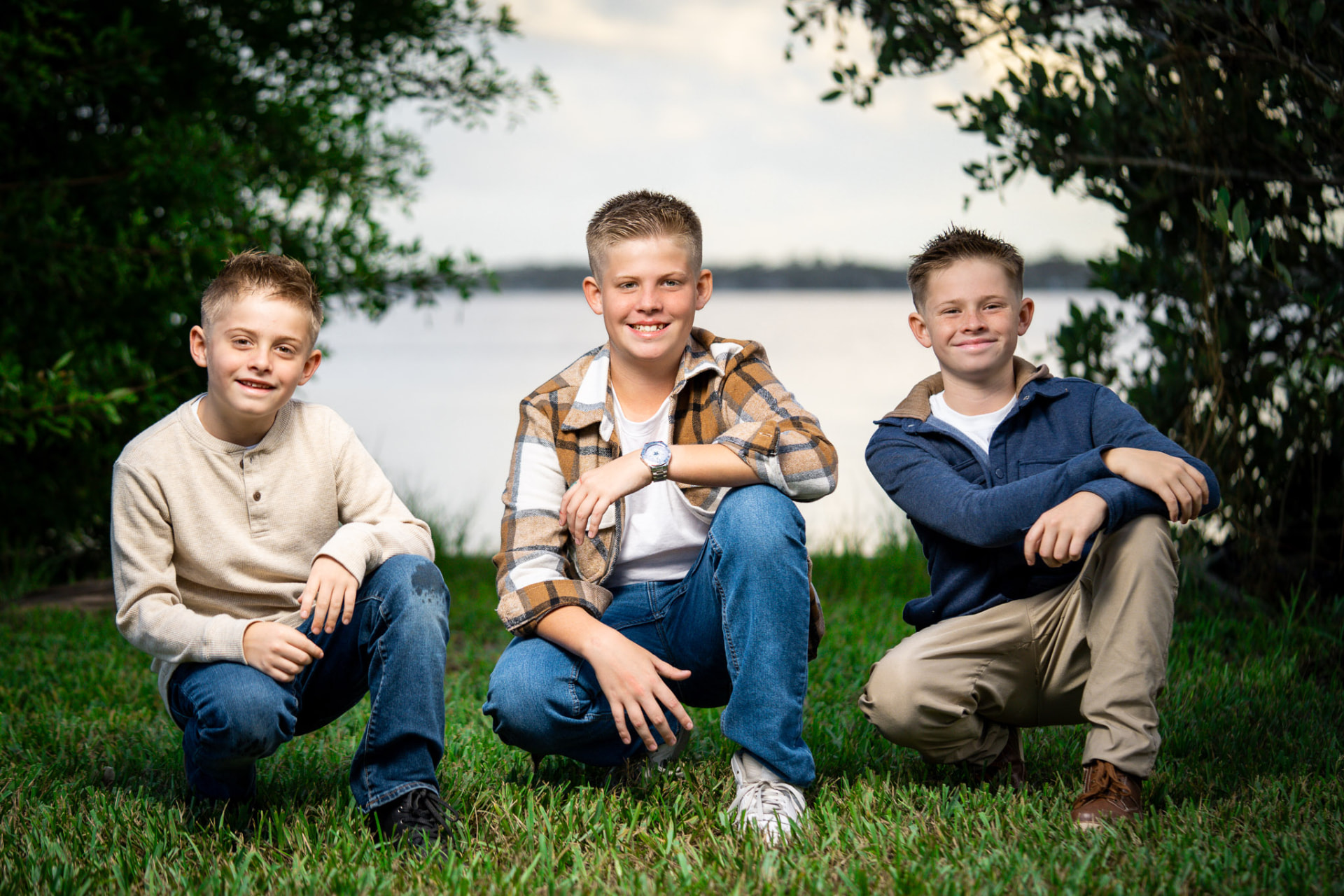 Three brothers crouching on green grass beside a lake, smiling beneath overhanging oak branches