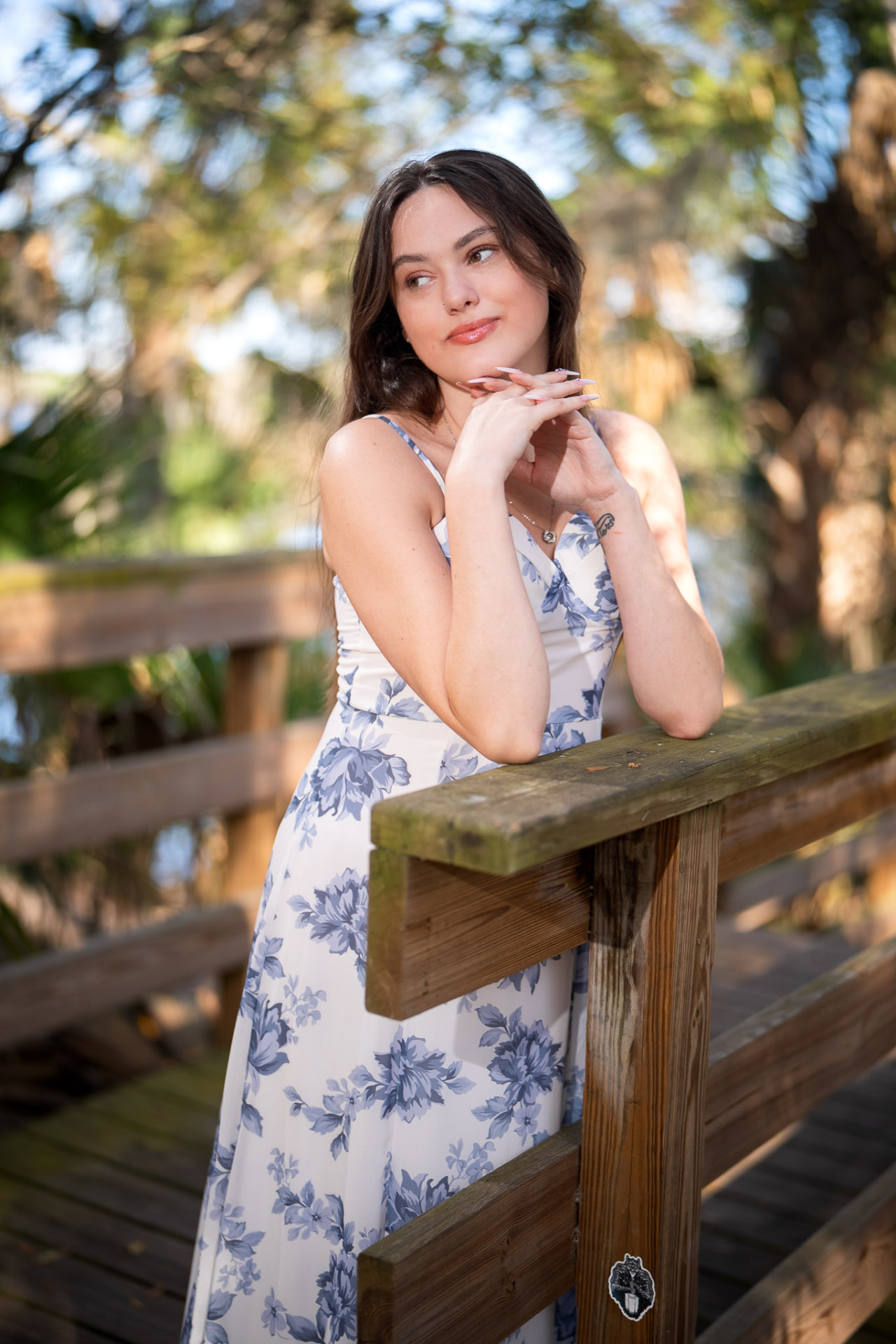 Young woman in a blue-and-white floral dress leaning on a wooden railing in dappled garden light
