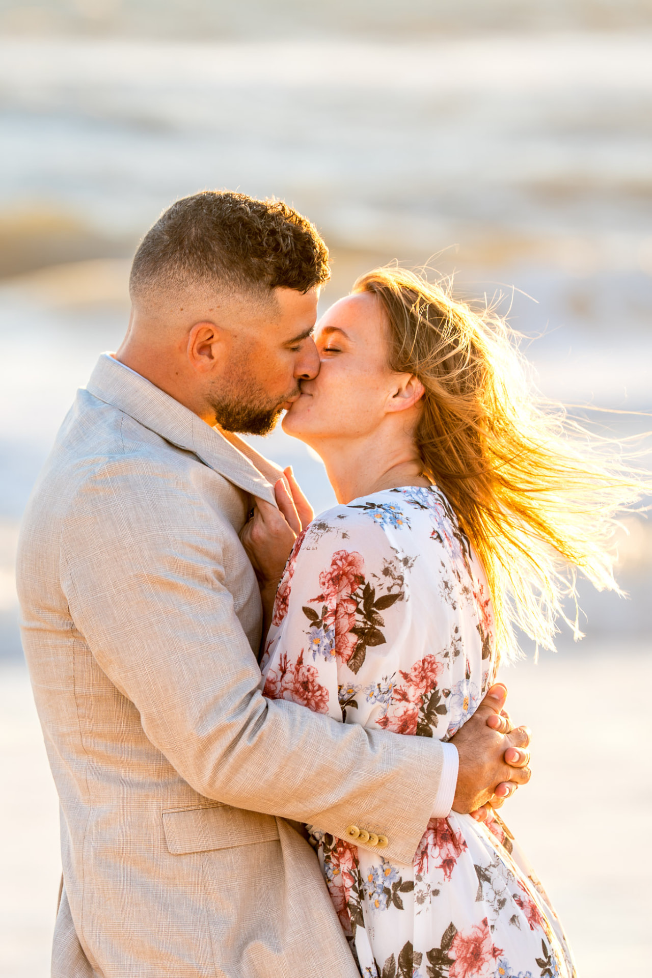 Couple sharing a kiss on the beach in golden light, her floral dress and windswept hair against the soft ocean horizon