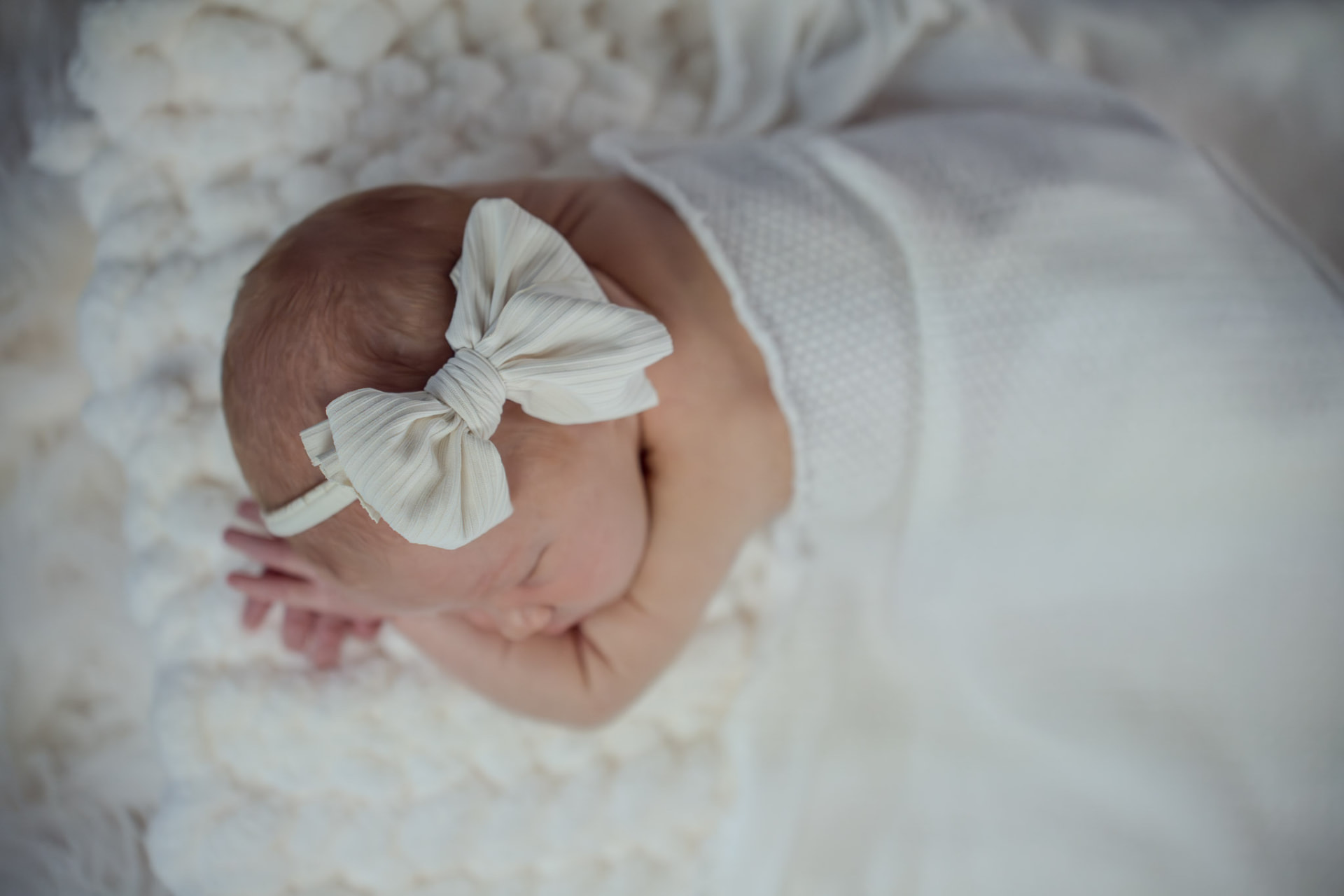 Sleeping newborn wearing a large cream bow, curled on a soft white textured blanket