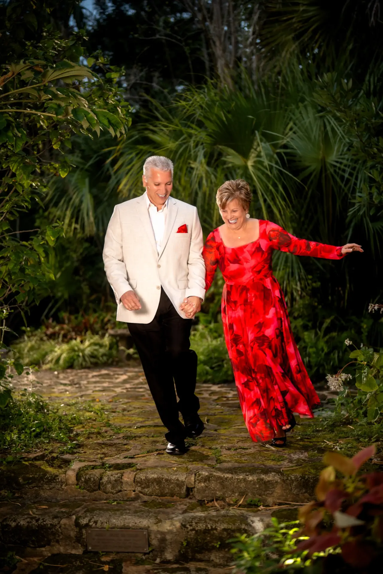 Couple holding hands and laughing as they walk up a mossy stone path surrounded by tropical palms and greenery