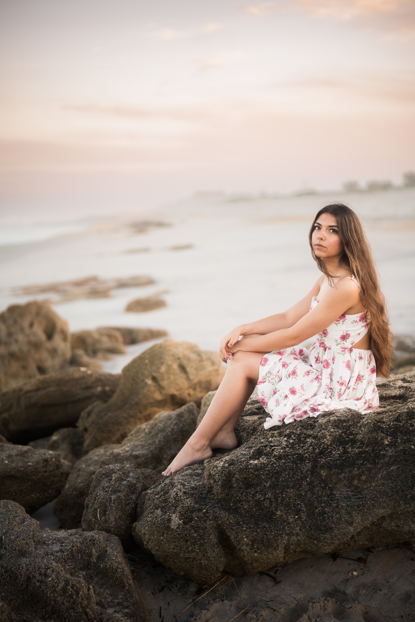 Senior portrait of a young woman in a floral dress seated on coquina rocks at the shoreline during a pink sunrise