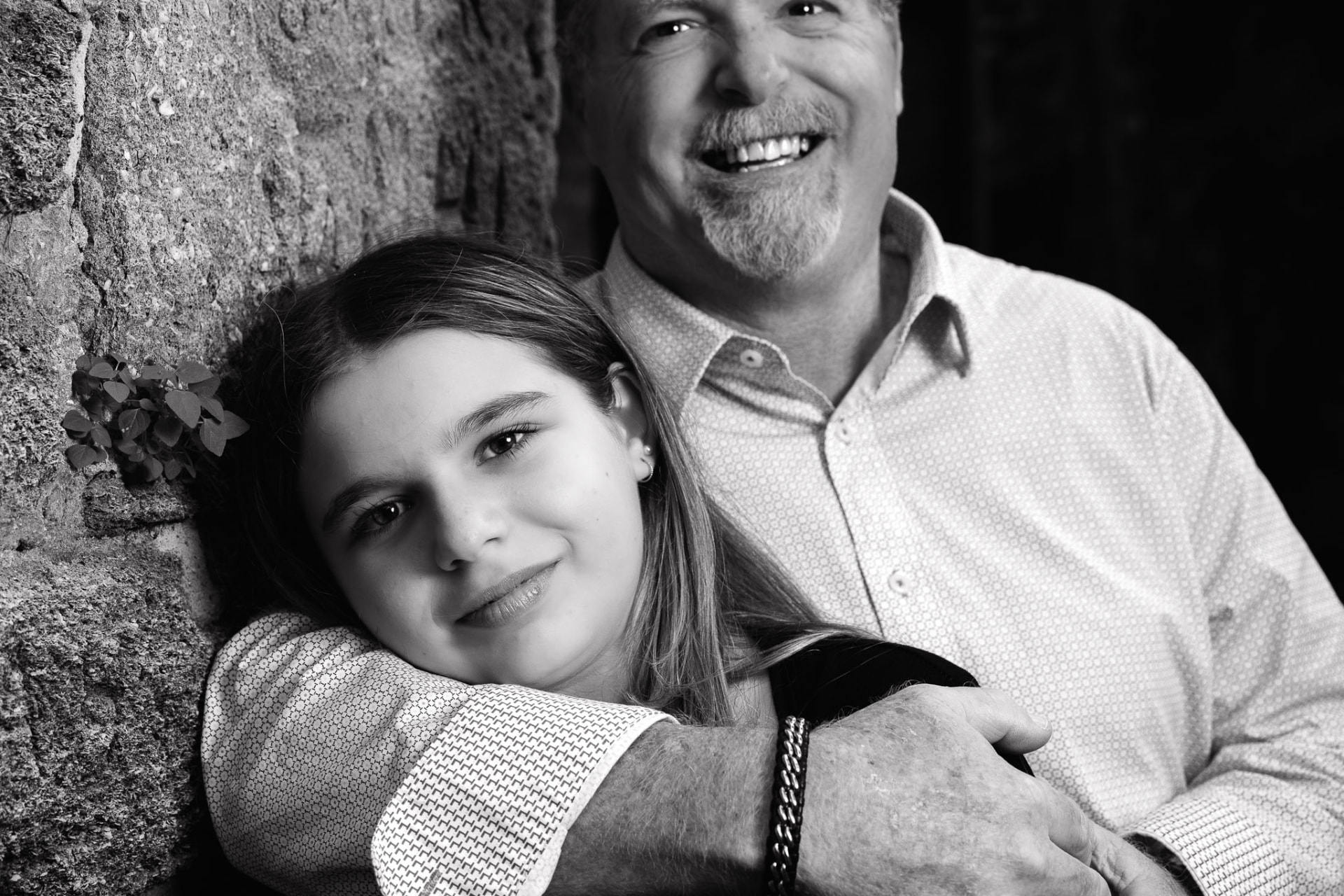 Black-and-white portrait of a father and daughter smiling together against a stone wall