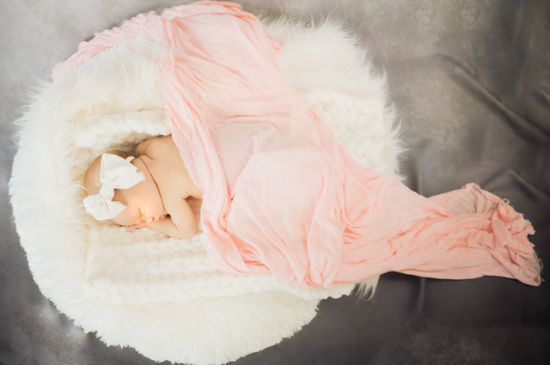 Sleeping newborn swaddled in a flowing pink wrap, nestled on a white fur prop