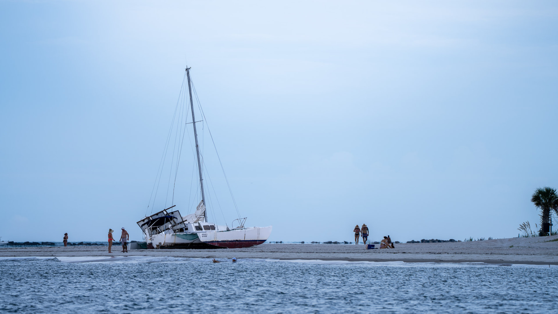 A beached sailboat resting on the sand at dusk with people gathered nearby and calm surf in the foreground
