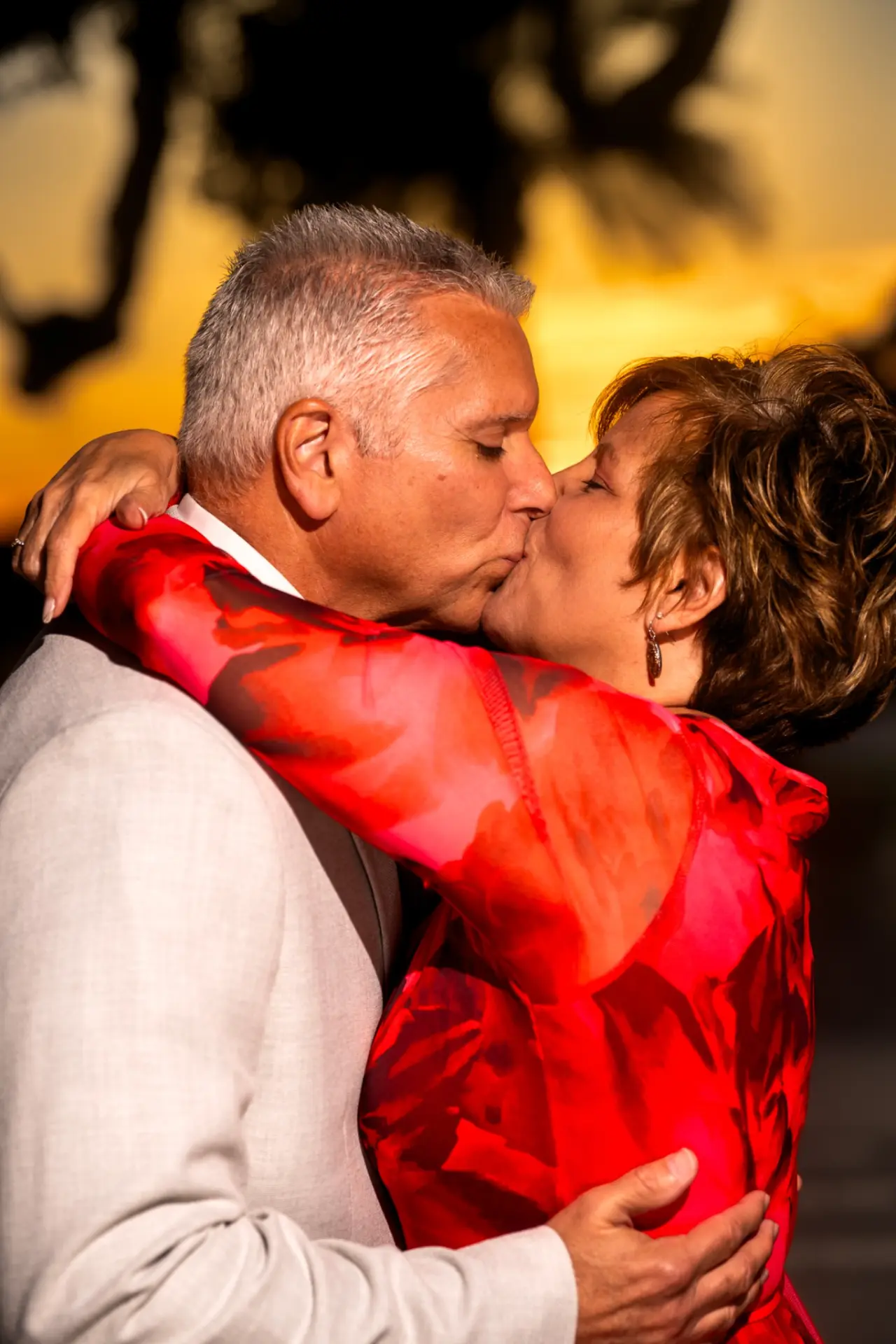 Close-up of a couple kissing in a warm embrace against a golden sunset backdrop, the woman's arm draped over his shoulder