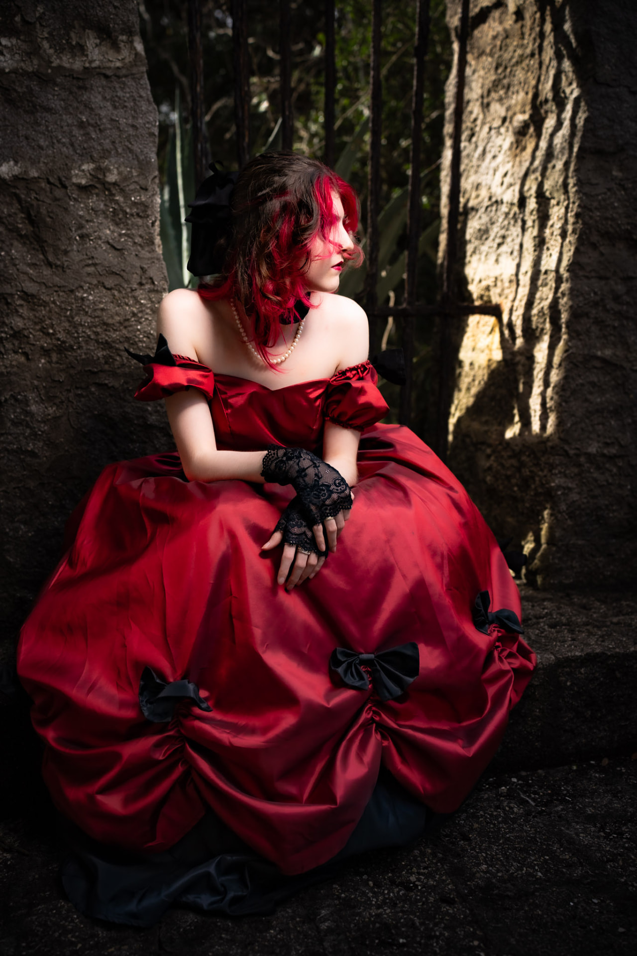 Woman in a red satin ball gown and black lace gloves seated on stone steps, looking away into the light