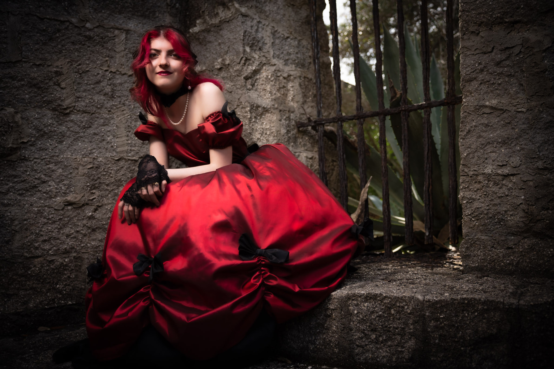 Woman with vivid red hair in a red satin ball gown with black bows seated against a weathered coquina wall beside an iron gate
